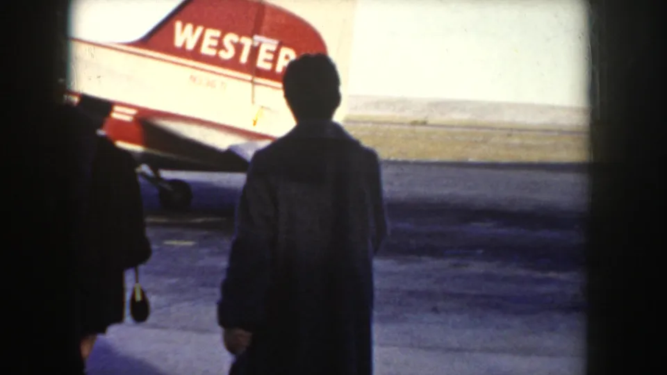 Western Airlines propeller aircraft on tarmac at what may be an Idaho airport, circa 1955; silhouetted passengers approach including a figure carrying a handbag; red tail livery clearly legible.