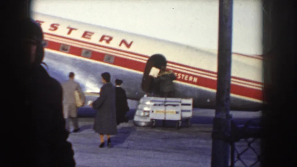 Western Airlines aircraft fuselage and red livery stripe, text partially obscured by a tarmac pole; three passengers approach the plane with a loaded baggage cart visible, circa 1955.