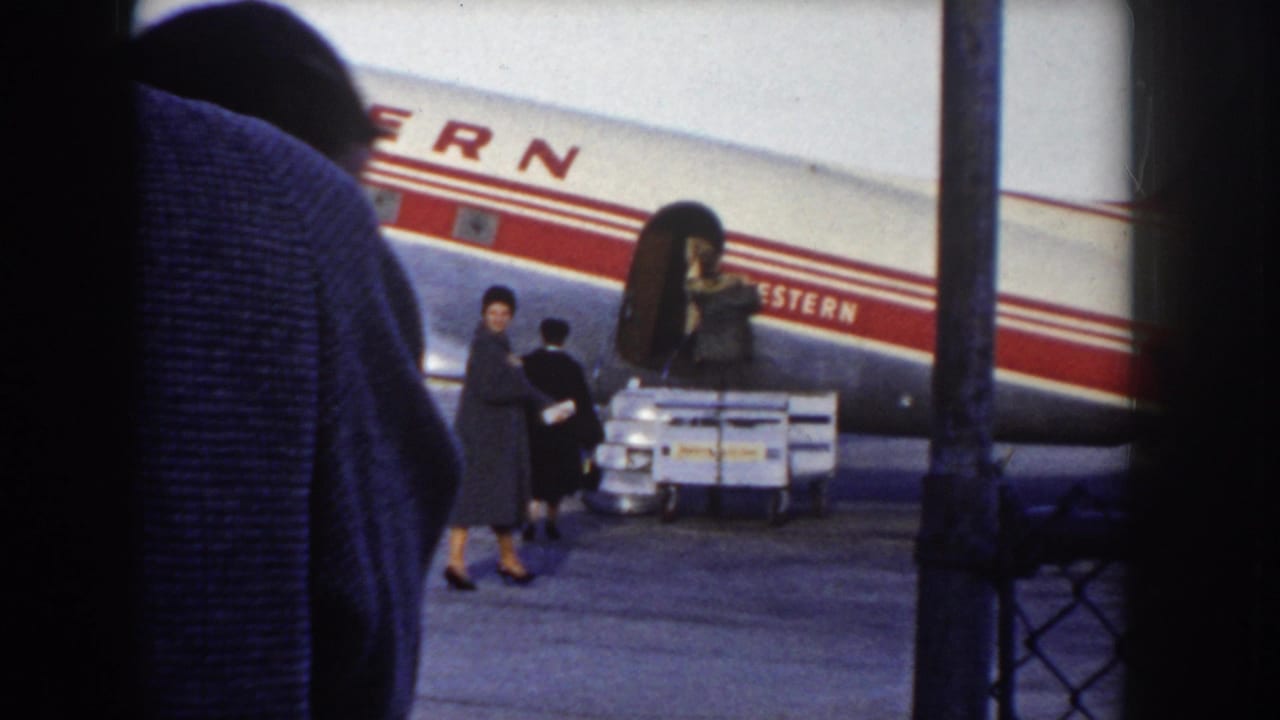8mm frame showing a woman in a dark coat and hat, a possible child, and a crate-laden luggage cart on the tarmac alongside a Western Airlines aircraft, c.1955.