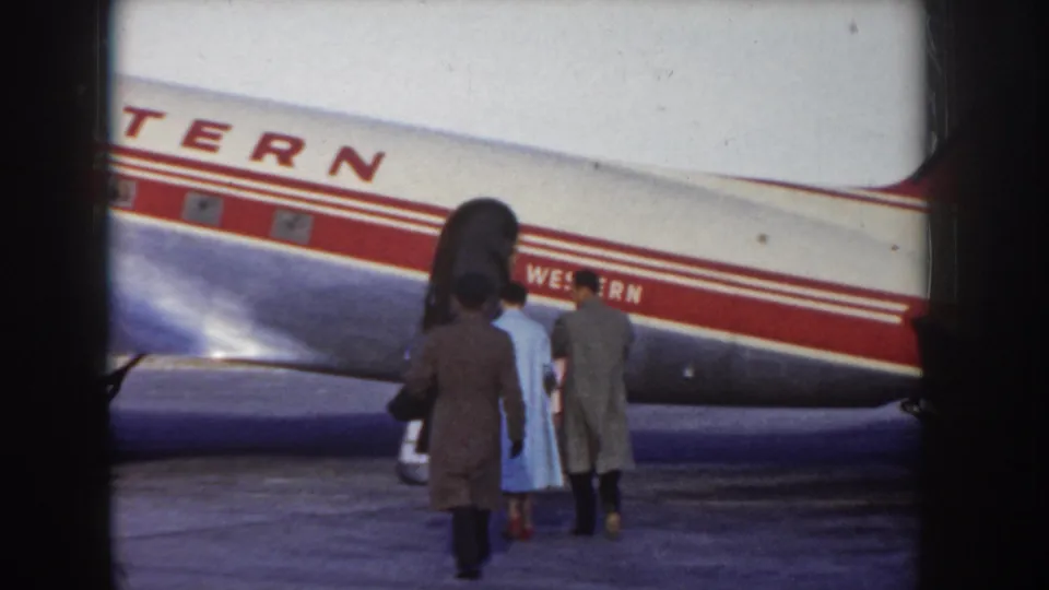 8mm frame showing figures in mid-century dress — including a light blue coat — alongside a Western Airlines aircraft during tarmac departure, Idaho USA, c.1955.