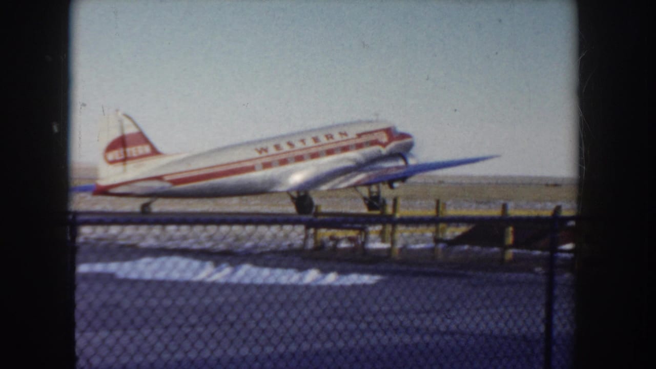 Western Airlines twin-engine propeller aircraft in red-and-white livery taxiing on a snowy Idaho airfield, circa 1955, shot through a chain-link perimeter fence on 8mm color film.
