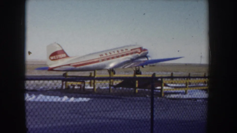 Western Airlines twin-engine aircraft advancing along tarmac at an Idaho airfield circa 1955, framed through chain-link fence with snow patches visible on the ground.