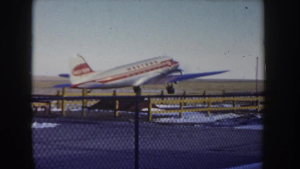Western Airlines propeller aircraft taxiing further along a snow-patched Idaho airfield circa 1955, viewed through chain-link fencing with yellow wooden posts prominent in foreground.