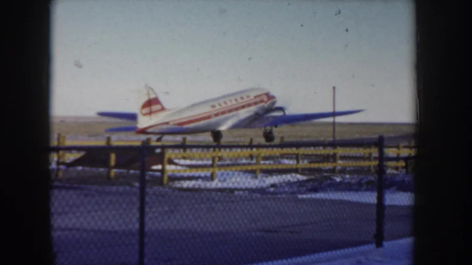 Western Airlines twin-engine propeller aircraft approaching the runway at an Idaho airfield circa 1955, chain-link fence and yellow posts in foreground, snow visible on the ground.