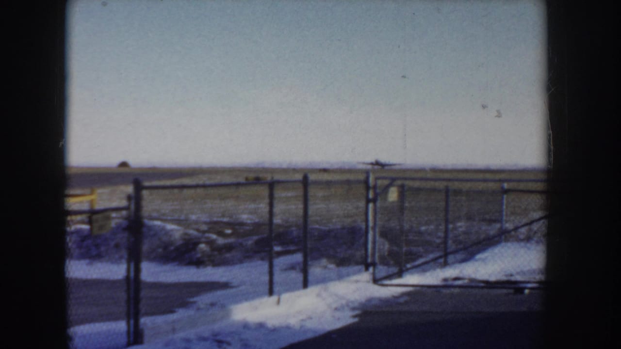 Chain-link perimeter fence at a small Idaho airfield, circa 1955, with snow banked along the base and a flat open tarmac visible behind it; 8mm home movie.