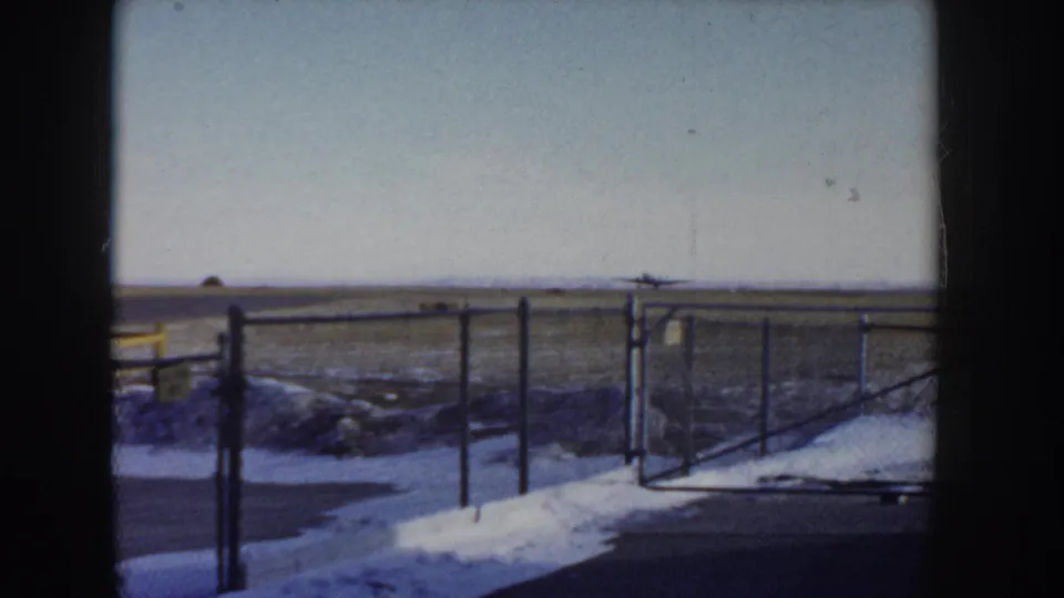 Chain-link perimeter fence at a small Idaho airfield, circa 1955, with snow banked along the base and a flat open tarmac visible behind it; 8mm home movie.