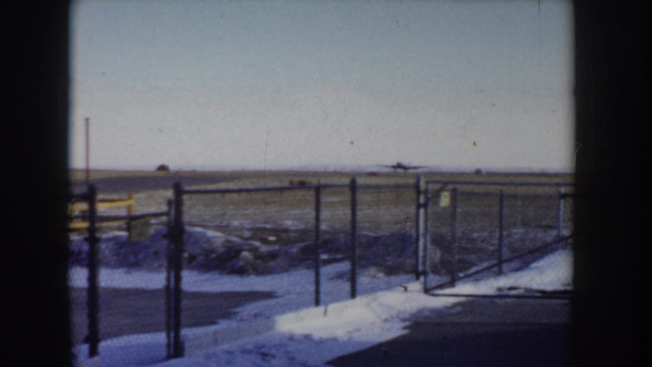 Gate section of chain-link perimeter fence at a small airfield, Idaho circa 1955, with snow on the ground and a flat open plain extending to the horizon.