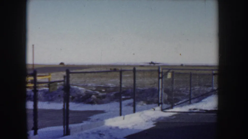 Gate section of chain-link perimeter fence at a small airfield, Idaho circa 1955, with snow on the ground and a flat open plain extending to the horizon.