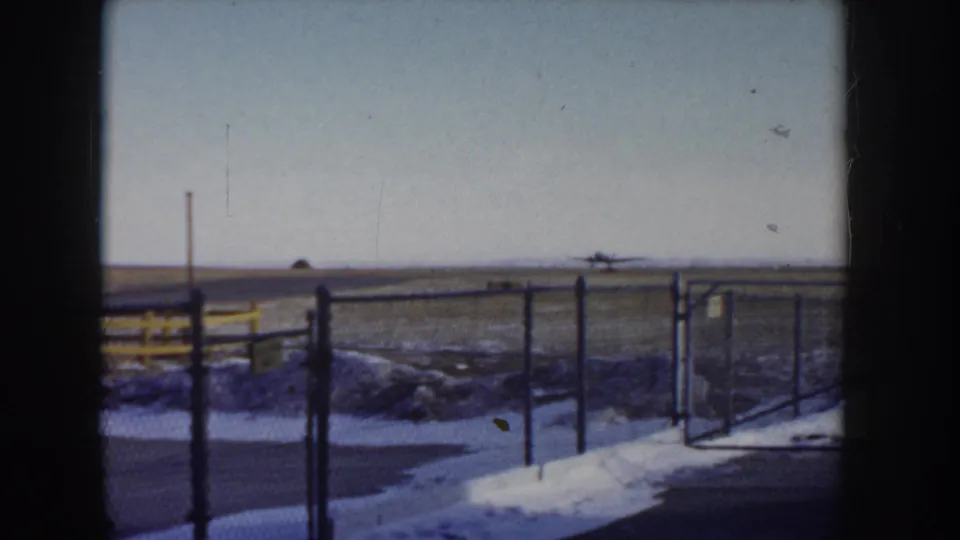 Ground-level view through chain-link fencing at a small rural airfield, Idaho circa 1955, with snow-covered verge and distant upright poles at the horizon.