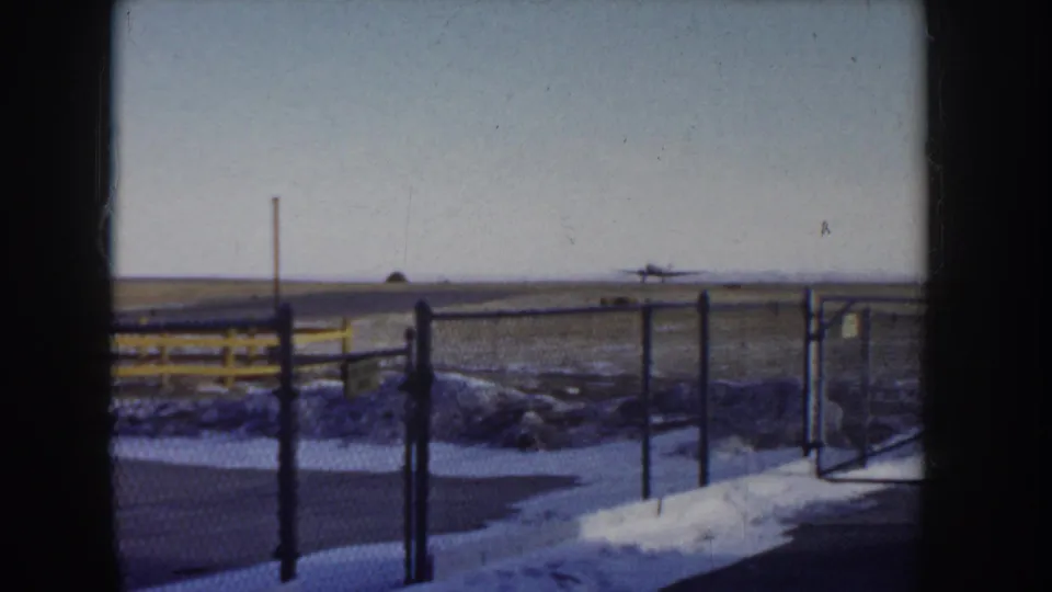 Near-static 8mm frame of chain-link fencing at a small Idaho airfield, circa 1955, with snow-edged tarmac verge and flat plain extending to the skyline.