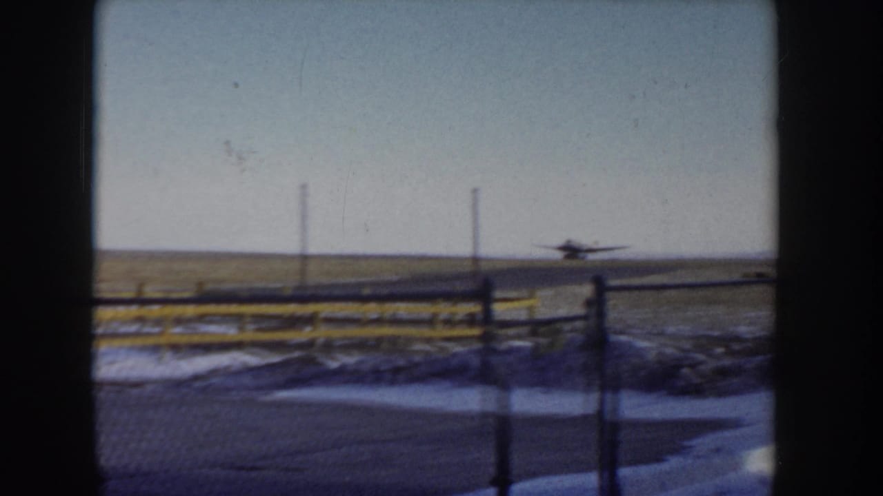 Small single-engine propeller aircraft on the runway at what may be a rural Idaho airfield, circa 1955, shot through a chain-link fence with yellow safety posts, snow-dusted ground.