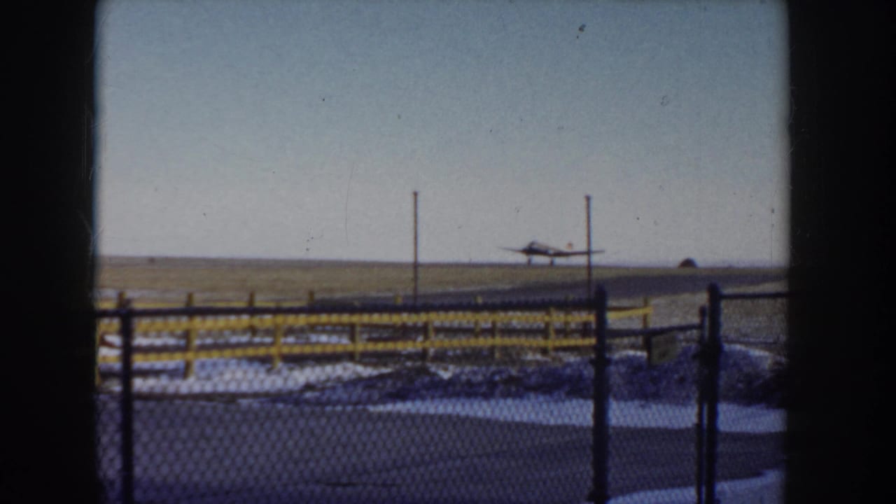 Small propeller plane advancing along the runway, chain-link fence with yellow posts in foreground, snow patches visible.