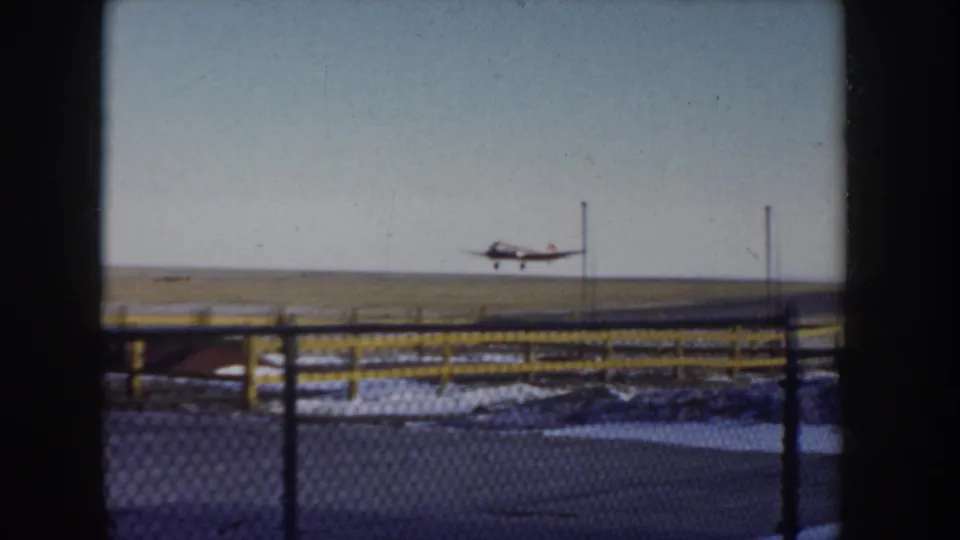 Small single-engine propeller aircraft just airborne at what may be a rural Idaho airfield, circa 1955; chain-link fence with yellow posts in foreground, snow-patched ground below.