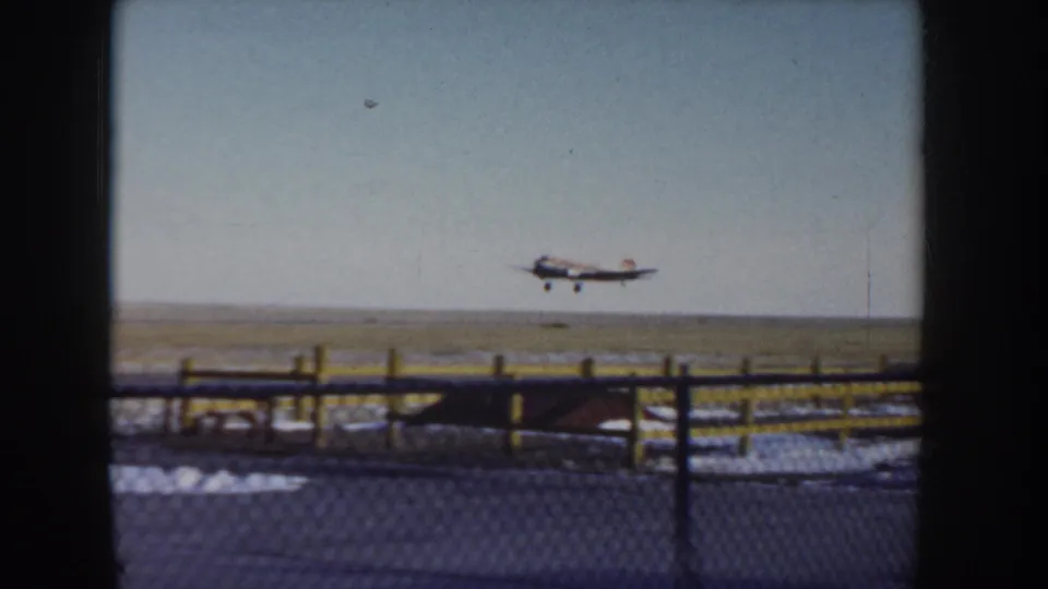Small single-engine propeller aircraft climbing away from what may be a rural Idaho airfield, circa 1955; chain-link perimeter fence and yellow posts in foreground, flat snow-dusted landscape.