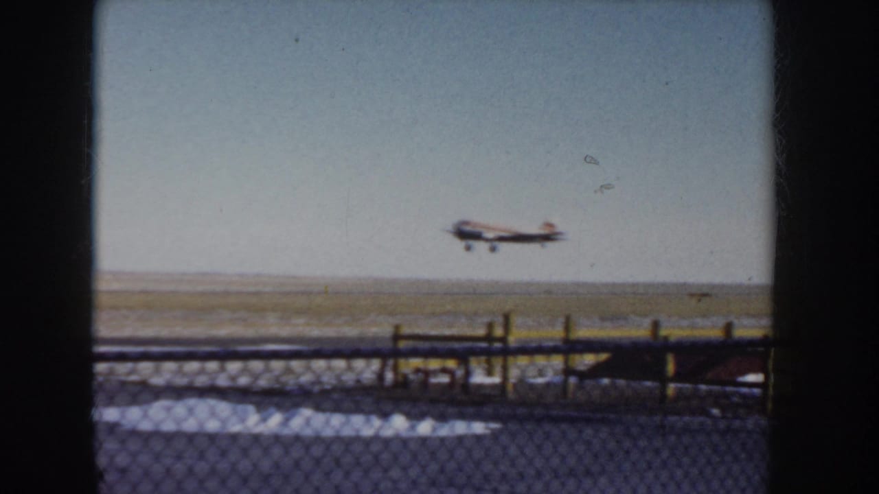8mm home-movie frame, c.1955 Idaho: twin-engine propeller plane airborne over flat open landscape, with chain-link fence in foreground.