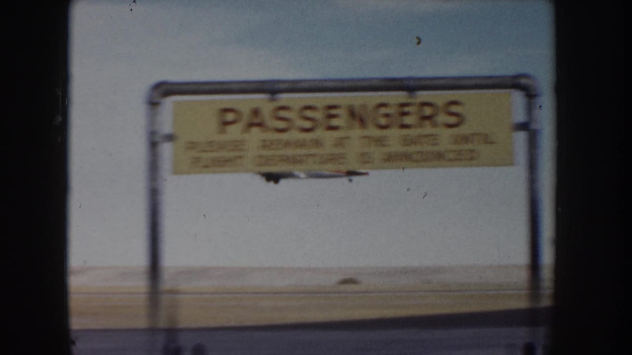 8mm frame showing a yellow-painted 'PASSENGERS' gate sign on a metal pipe frame at what appears to be a small mid-1950s airfield, c.1955.