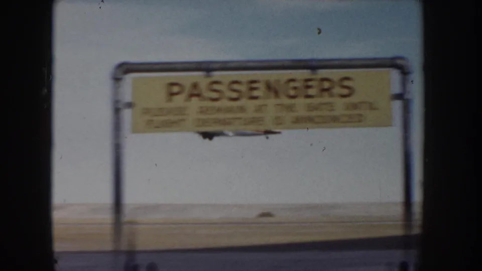 8mm frame showing a yellow-painted 'PASSENGERS' gate sign on a metal pipe frame at what appears to be a small mid-1950s airfield, c.1955.