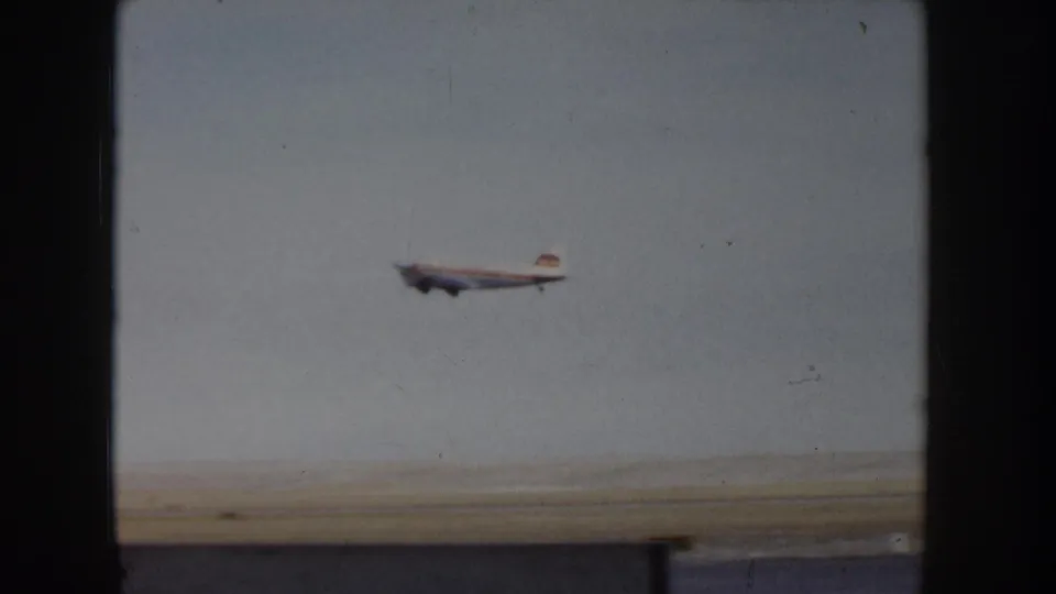 8mm home-movie frame, c.1955: small twin-engine propeller aircraft at low altitude climbing against a pale sky, likely shortly after takeoff from a rural Idaho airfield.