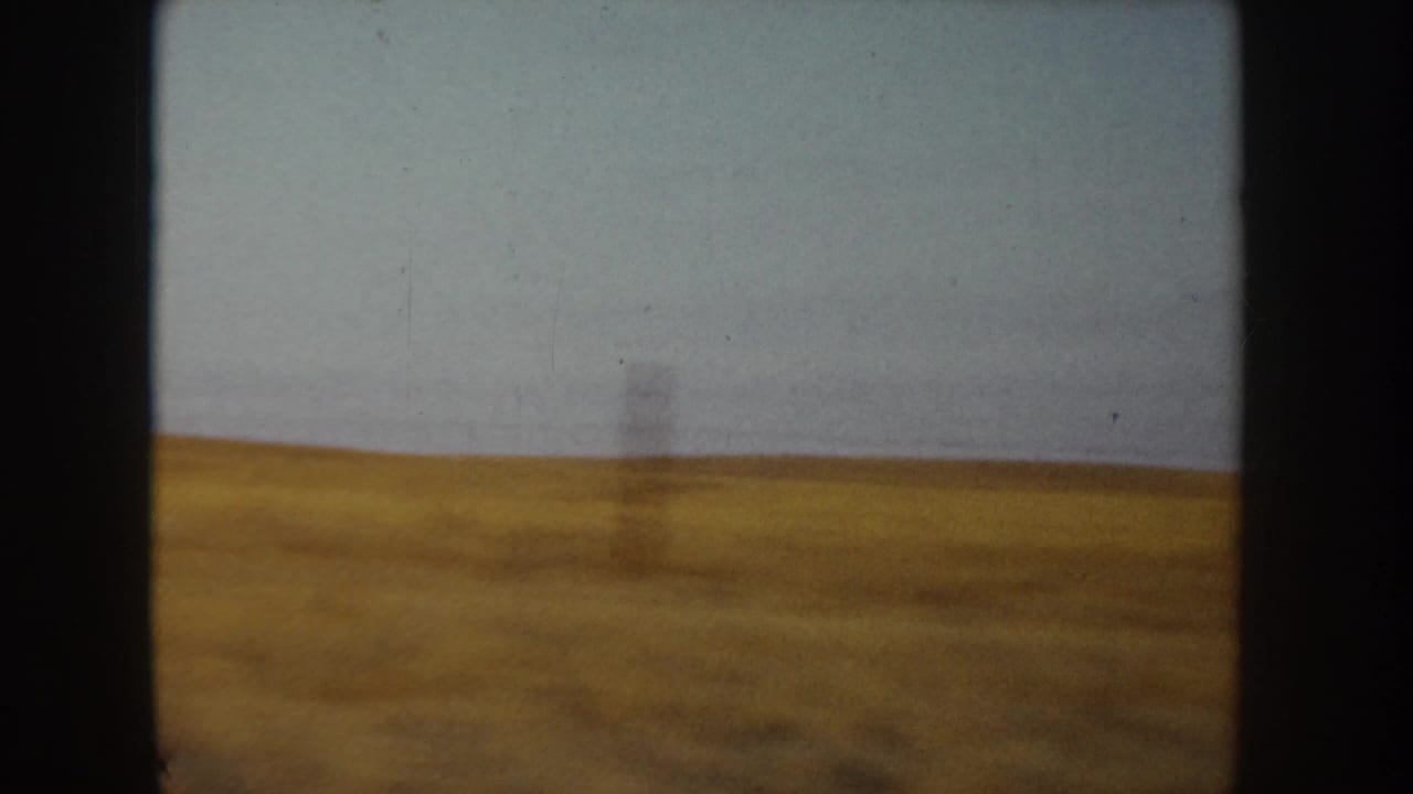 8mm home-movie frame, Idaho c.1955 — a dry golden field stretches to the horizon under a pale overcast sky, with a slender vertical element at mid-field.