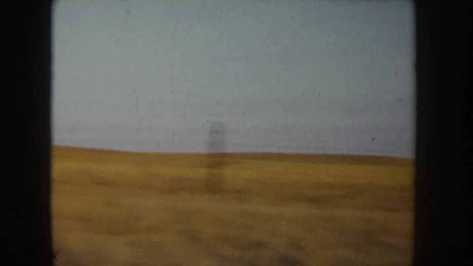 8mm home-movie frame, Idaho c.1955 — a dry golden field stretches to the horizon under a pale overcast sky, with a slender vertical element at mid-field.