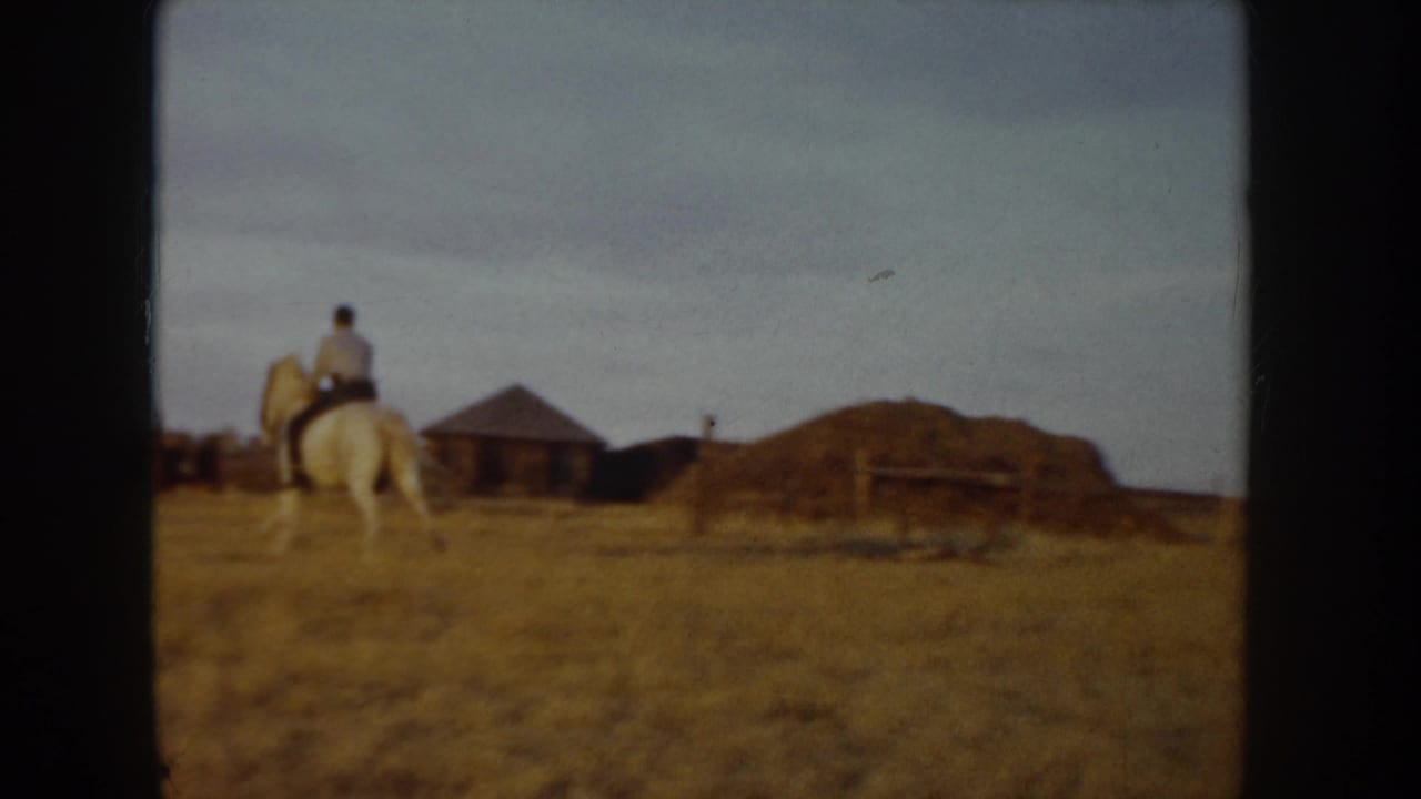 Rider on a white horse seen from behind, passing farm outbuildings and an earth mound across a dry golden field.