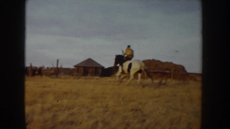 8mm home-movie frame, Idaho farm c.1955 — rider on white horse crosses dry grass left to right; low outbuilding and earth mound sit beneath an overcast sky.