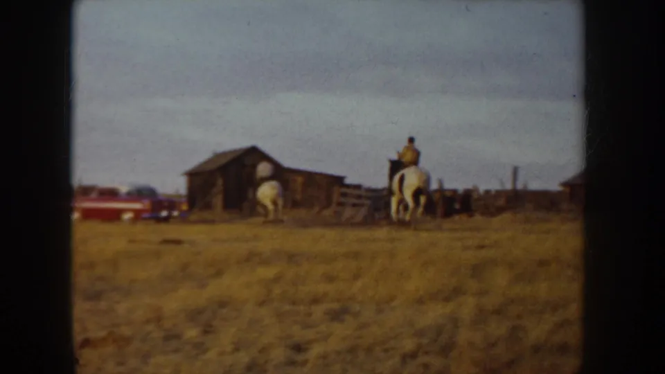 8mm home-movie frame, Idaho farm c.1955 — rider on white horse moves past a dark timber barn with a red period vehicle parked alongside; wooden fencing visible in the background.