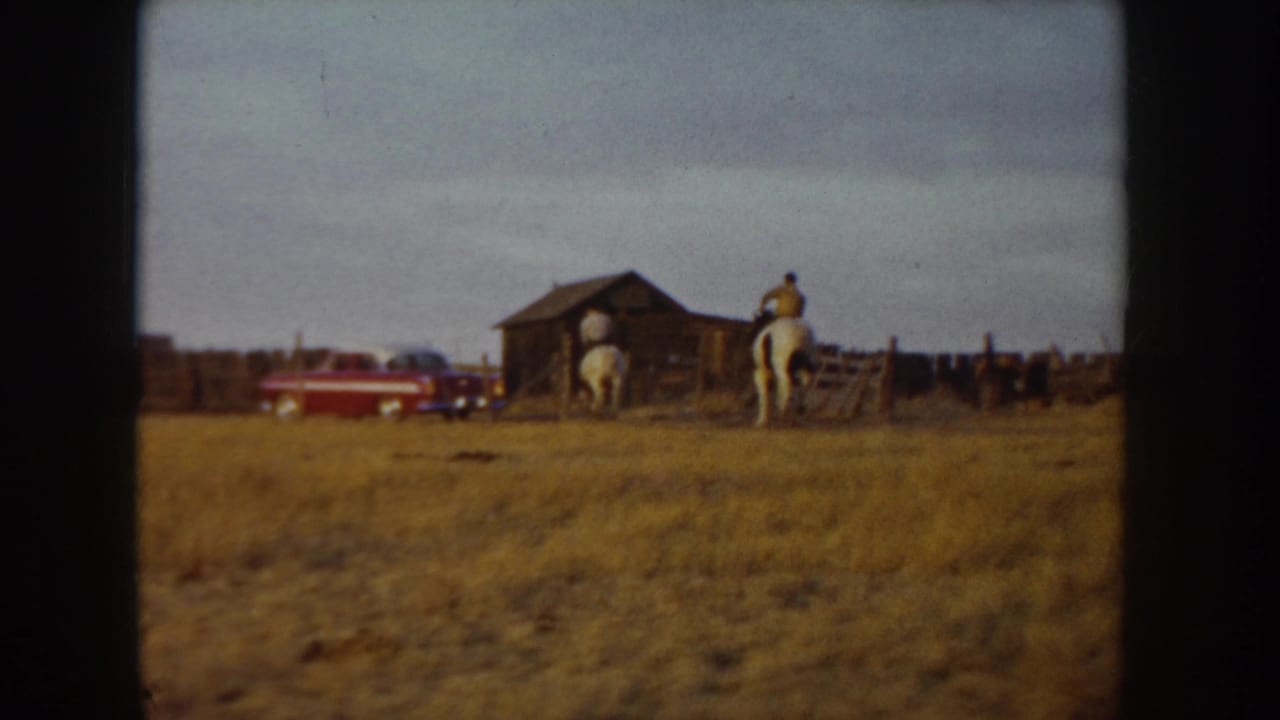 8mm home movie frame, Idaho ca. 1955: horse and rider moving toward a rustic barn, red mid-century automobile parked at left, golden open range in foreground.