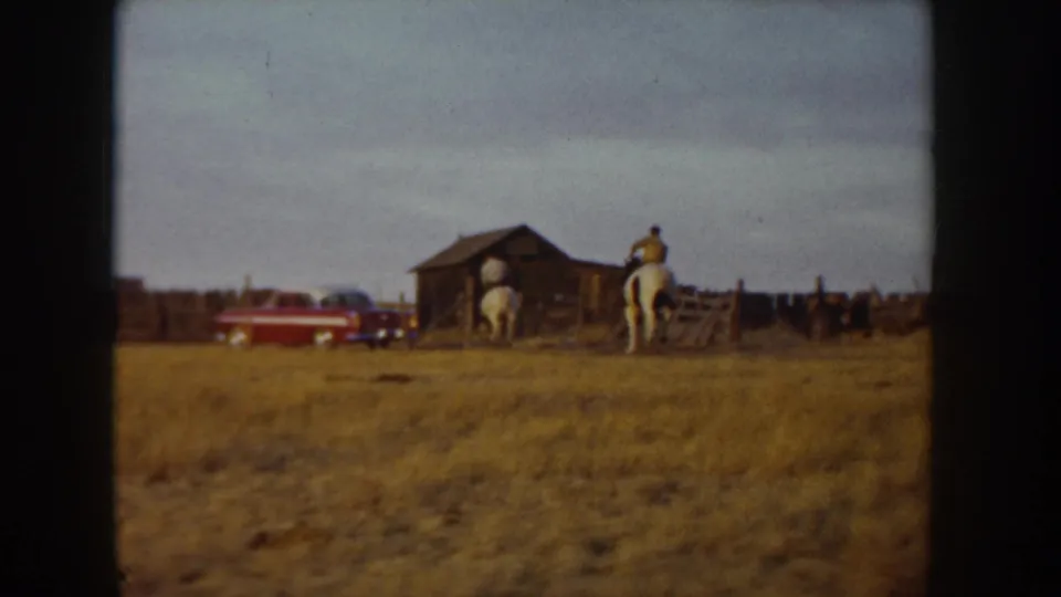 8mm home movie frame, Idaho ca. 1955: horse and rider moving toward a rustic barn, red mid-century automobile parked at left, golden open range in foreground.