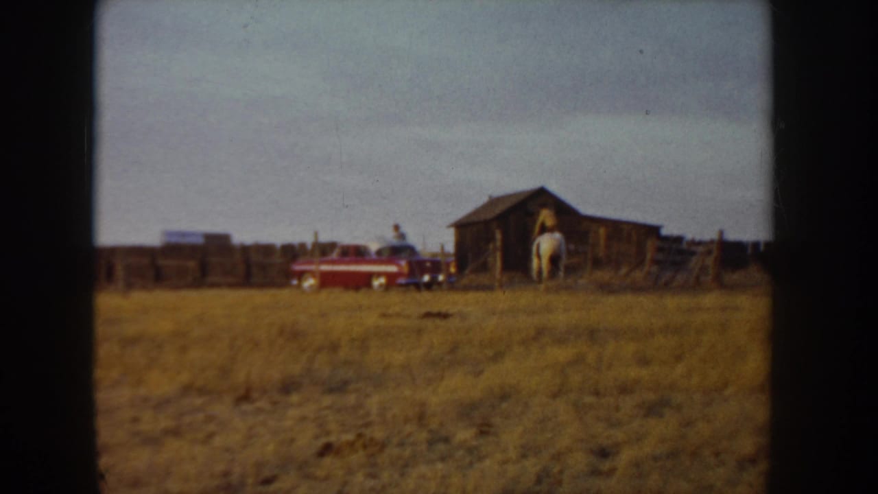 Wide shot of a dry grass field with a wooden barn, red vehicle, and a horse and rider visible in the mid-ground.