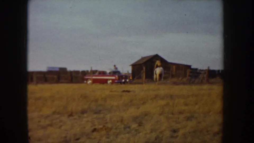 8mm home movie frame, Idaho ca. 1955: horse and rider near a rustic barn, red automobile parked at right, golden open range in foreground, second figure visible near barn.