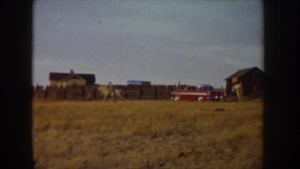 8mm home movie frame, Idaho ca. 1955: wide-angle view of a ranch complex with multiple wooden structures, red automobile, and horses with riders visible at distance.