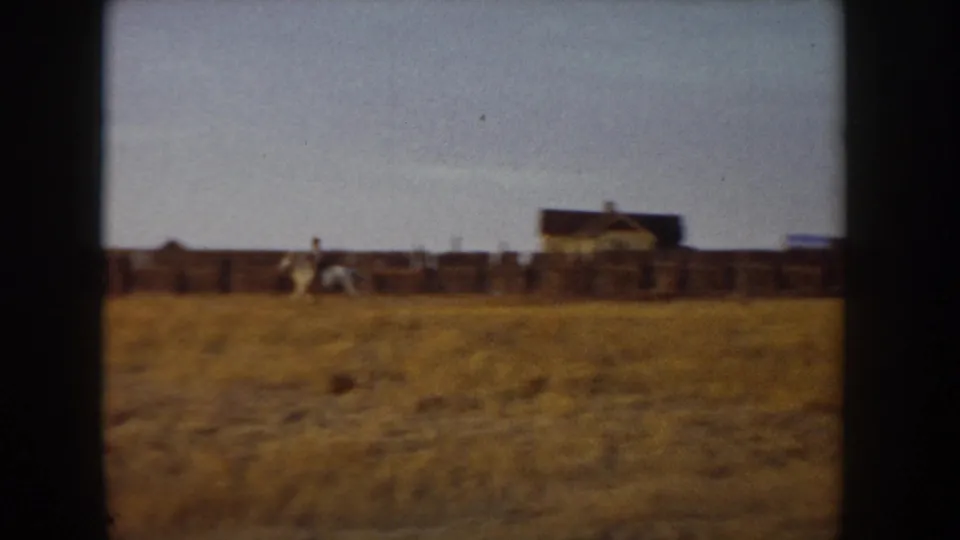 8mm home movie frame, Idaho ca. 1955: ranch outbuildings and corrals with a horse and rider near the barn, large expanse of golden dry grassland in foreground.