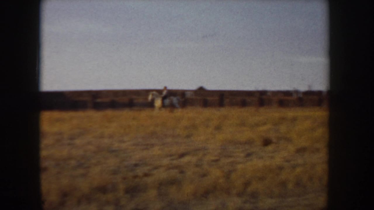 Wide 8mm shot of a rider on a light-colored horse in an open golden field with wooden post-and-rail fencing, Idaho, circa 1955.