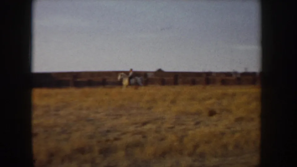 Wide 8mm shot of a rider on a light-colored horse in an open golden field with wooden post-and-rail fencing, Idaho, circa 1955.