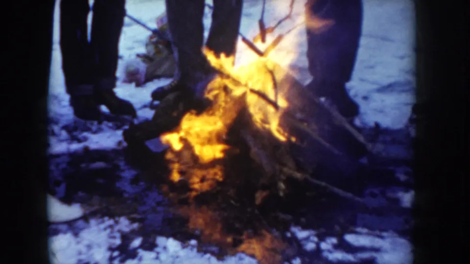 Tight 8mm shot of a group around an active campfire in snowy conditions; lower bodies only visible, dark trousers and white footwear, Idaho, circa 1955.