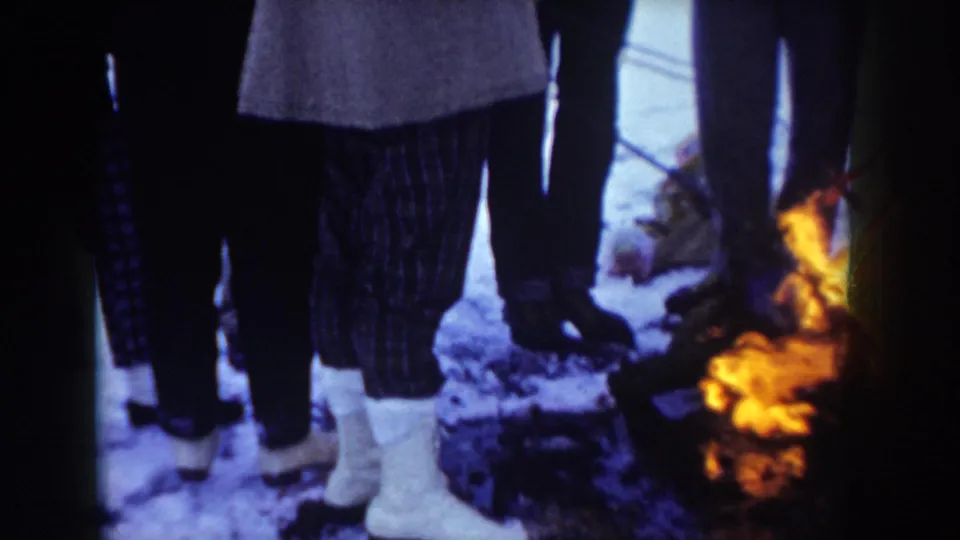 8mm close shot of a group gathered around a campfire in winter, lower bodies only; one person in light-colored trousers and white ankle boots, Idaho, circa 1955.