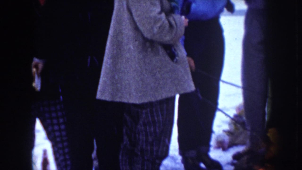 Winter-clad adults outdoors in snow, possibly Idaho circa 1955; close-up 8mm framing emphasizes layered cold-weather clothing; small figure crouches in background snow.