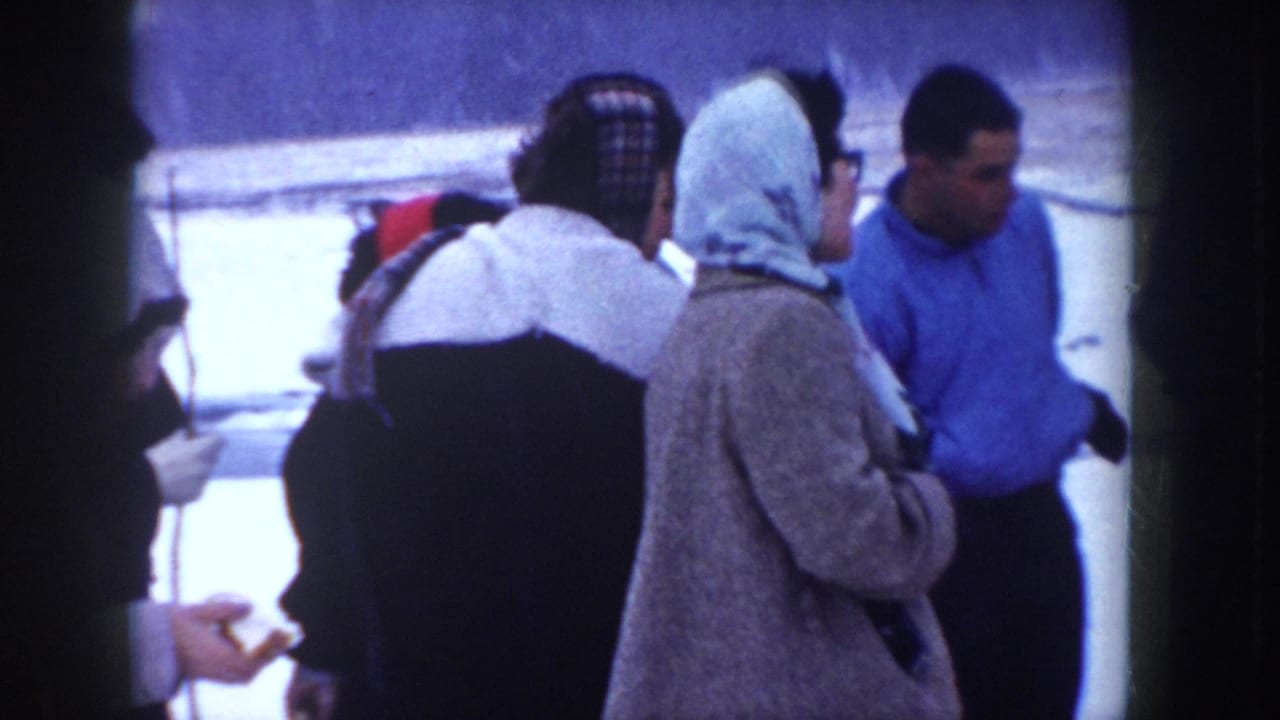 Group of adults seen from behind and in profile stand together in snow; women wear headscarves; one person holds a small object.