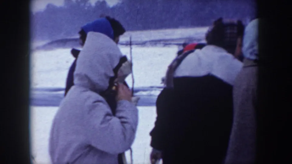 Close-up of two adults in winter outerwear standing in snow, possibly Idaho circa 1955; left figure in hooded coat appears to eat or drink; 8mm color grain evident.