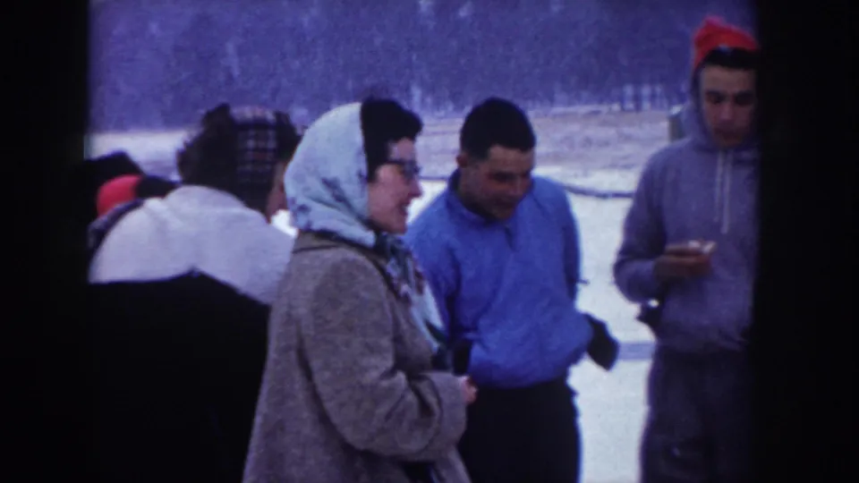 Adults in 1950s winter dress gathered outdoors in snow, possibly Idaho circa 1955; woman in headscarf and glasses stands with a man in a blue sweatshirt and another in a red knit hat.