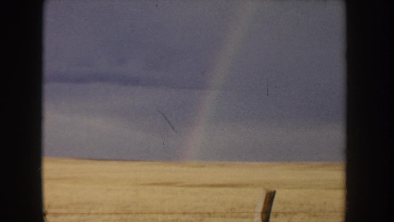 8mm home movie, Idaho c.1955 — golden grain field in lower frame, overcast blue-gray sky above, faint rainbow arc visible at left, wooden fence post standing in the field.