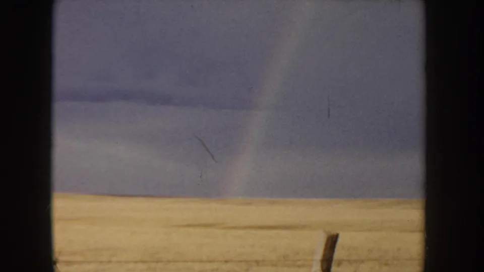 8mm home movie, Idaho c.1955 — golden grain field in lower frame, overcast blue-gray sky above, faint rainbow arc visible at left, wooden fence post standing in the field.