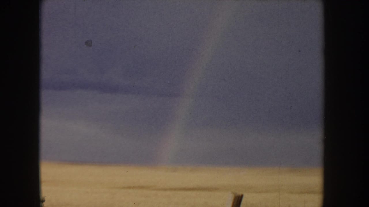 Golden grain field and overcast sky with a faint rainbow arc at right edge and a wooden fence post, 8mm home movie c.1955.