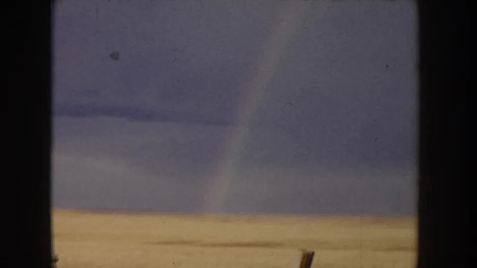 8mm home movie, Idaho c.1955 — golden grain field band across lower frame, pale overcast sky, faint rainbow arc at right edge, wooden fence post visible in the field.