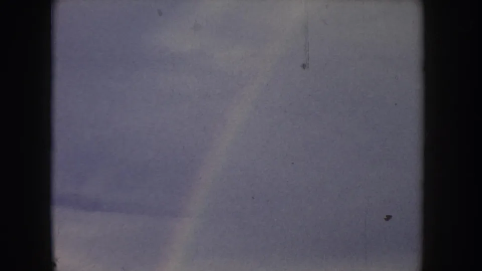 8mm home movie, Idaho c.1955 — overcast sky dominates the frame with a faint possible rainbow arc, characteristic grain, light scratches, and dust specks throughout.