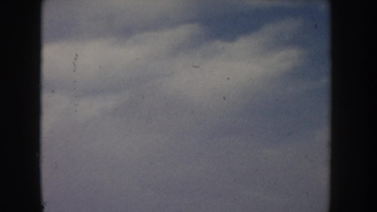 8mm home movie frame circa 1955, likely Idaho, showing a dramatic overcast sky with billowing grey-blue clouds and a sliver of flat golden grassland at the lower edge.