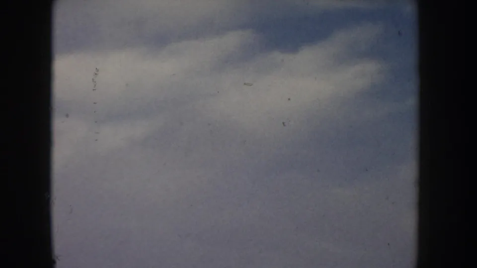 8mm home movie frame circa 1955, likely Idaho, showing a dramatic overcast sky with billowing grey-blue clouds and a sliver of flat golden grassland at the lower edge.
