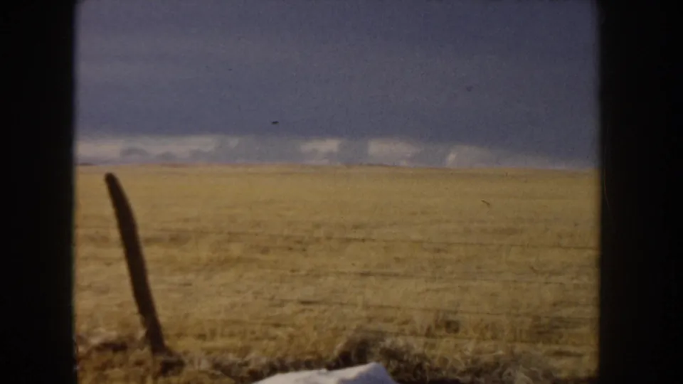 8mm home movie frame, circa 1955, likely Idaho, showing expansive flat grassland under a stormy overcast sky with a weathered leaning fence post at left.