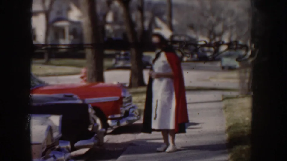 8mm color home movie frame, circa 1955 Idaho, showing a woman in white and a red cape beside a mid-century red automobile on a tree-lined suburban street with a white clapboard house.